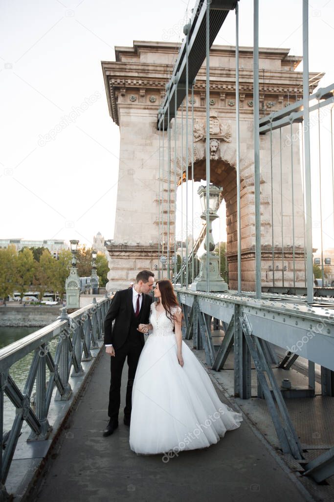 Bride and groom kissing in the old town street. Wedding couple walking on Szechenyi Chain Bridge, Hungary. Happy romantic young couple celebrating their marriage. Wedding and love concept.