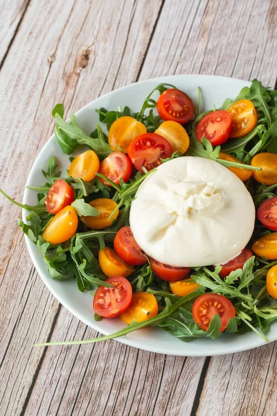Lezzetli, mozzarella burrata, roka, kiraz domatesleri ve ahşap bir arka planda beyaz bir tabakta fesleğen. Yaklaş. Sağlıklı İtalyan yemekleri
