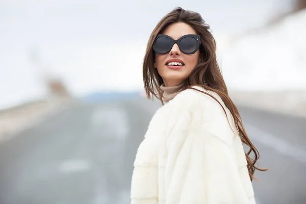 Menina posando na estrada no fundo floresta de inverno. Glamoroso engraçado jovem mulher com sorriso vestindo camisola elegante, casaco de pele elegante branco e óculos de sol. Pele e conceito de moda. Pessoas bonitas . — Fotografia de Stock