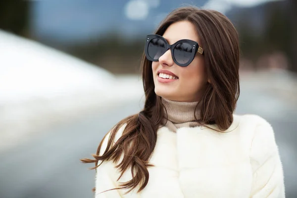 Menina posando na estrada no fundo floresta de inverno. Glamoroso engraçado jovem mulher com sorriso vestindo camisola elegante, casaco de pele elegante branco e óculos de sol. Pele e conceito de moda. Pessoas bonitas . — Fotografia de Stock