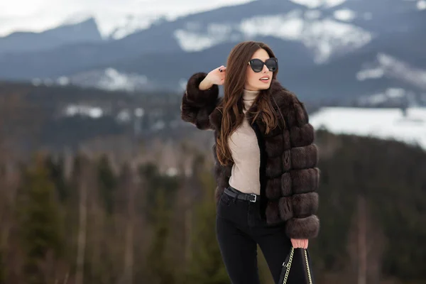 Flicka poserar på vintern berg bakgrund. Glamorös ung kvinna bär snygg tröja, mörka jeans, brun fashionabla kort pälsjacka och svart väska. Päls och mode koncept. Vackra människor. — Stockfoto