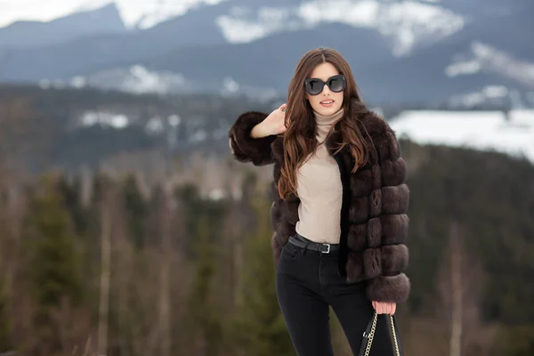 Dziewczyna pozowanie na tle zimowych gór. Czarująca młoda kobieta w stylowym swetrze, ciemnych dżinsach, brązowej, modnej krótkiej kurtce futerkowej i czarnej torbie. Futro i koncepcja mody. Piękni ludzie.. — Zdjęcie stockowe