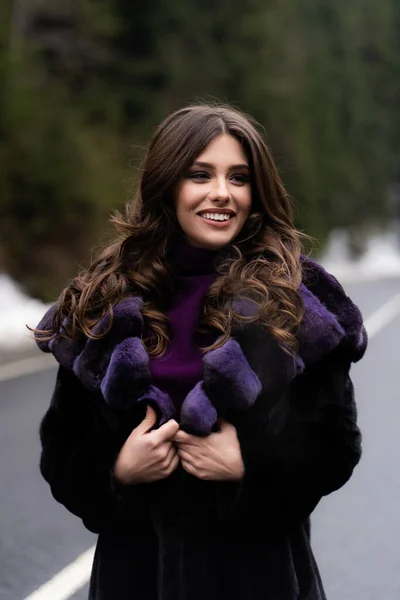 Κορίτσι χαμογελά στην κάμερα στο φόντο δάσος χειμώνα. Λαμπερό αστείο νεαρή γυναίκα με χαμόγελο φορώντας κομψό πουλόβερ, μωβ μοντέρνα γούνα παλτό. Γούνα και μόδα έννοια. Όμορφοι άνθρωποι. — Φωτογραφία Αρχείου