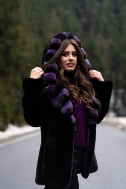 Kış ormanı arka planında kameraya poz veren kız. Gülümseyen, şık bir süveter giyen, mor moda kürklü, çekici, komik bir genç kadın. Kürk ve moda konsepti. Güzel insanlar..