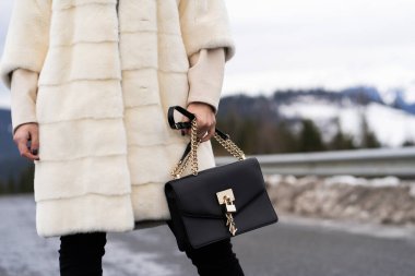 Şık bir kıyafet. Koyu renkli büyük siyah çantaya yaklaş. Sokakta poz veren, beyaz moda kürk giyen bir manken. Kürk ve moda konsepti. Güzel insanlar. Kadın moda konsepti.