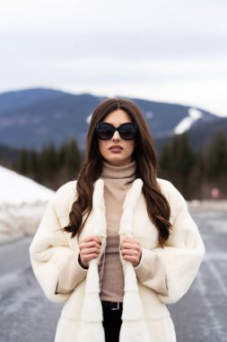 Kışın ormanda poz veren bir kız. Süslü süveter, beyaz moda kürk manto ve güneş gözlüğü takan alımlı, komik, genç bir kadın. Kürk ve moda konsepti. Güzel insanlar..