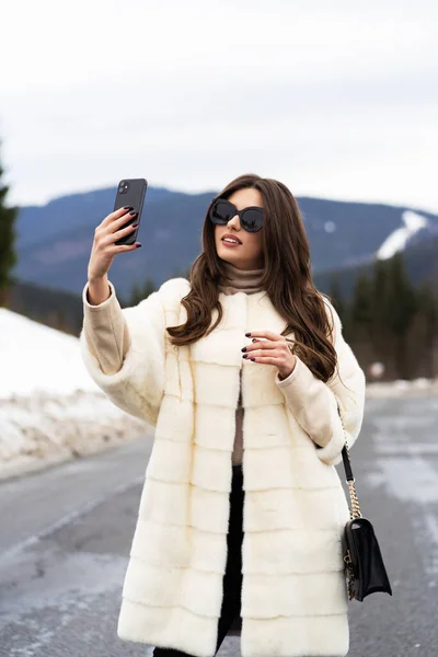 겨울 산에서 스마트폰으로 자신의 사진을 찍는 소녀. 멋진 스웨터를 입고, 하얀 모피 코트와 선글라스를 착용 한 젊은 여성. 패션 과 모피 개념. 아름다운 사람들. — 스톡 사진