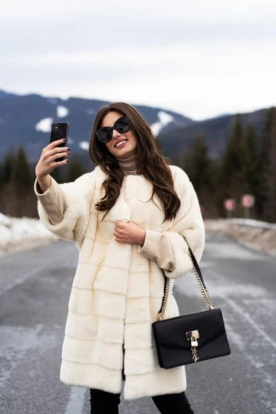 Дівчина фотографує себе на смартфоні на фоні зимових гір. Молода жінка в стильному светрі, білому модному шубі та сонцезахисних окулярах. Концепція хутра та моди. Прекрасні люди . — стокове фото