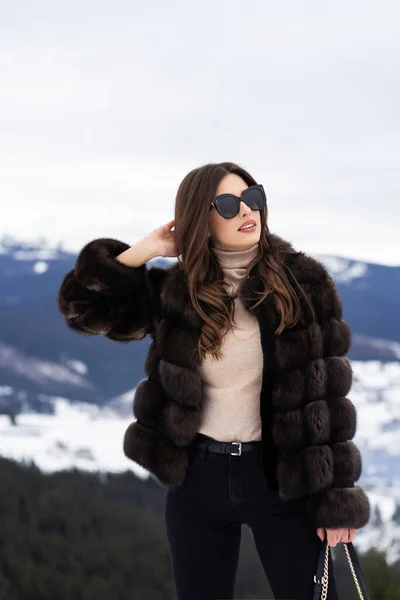 Girl posing on winter mountains background. Glamorous young woman wearing stylish sweater, brown fashionable short fur jacket and black bag and sunglasses. Fur and fashion concept. Beautiful people. — Stock Photo, Image