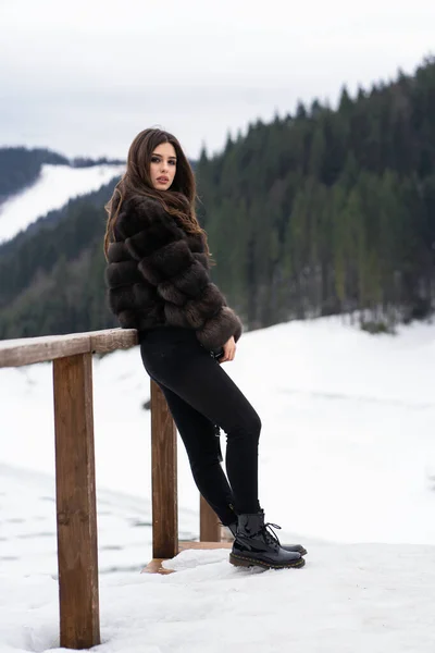 Κορίτσι που ποζάρει σε χειμερινά βουνά φόντο. Λαμπερή νεαρή γυναίκα που φοράει κομψό πουλόβερ, σκούρο τζιν, καφέ μοντέρνο κοντό γούνινο μπουφάν και μαύρη τσάντα. Γούνα και μόδα έννοια. Όμορφοι άνθρωποι. — Φωτογραφία Αρχείου
