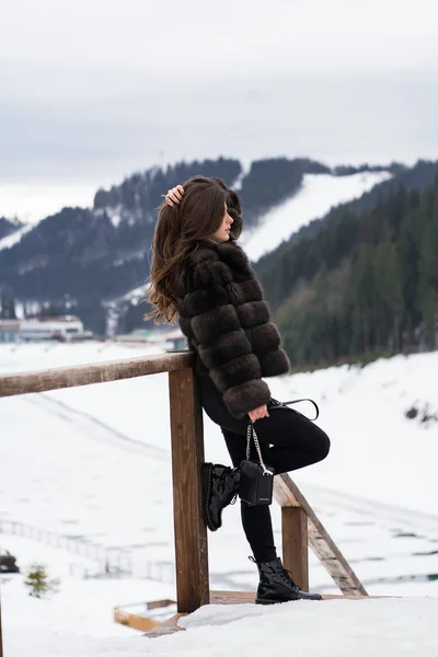 Flicka poserar med upphöjd arm på vintern berg bakgrund. Glamorös ung kvinna bär snygg tröja, mörka jeans, brun fashionabla kort pälsjacka. Päls och mode koncept. Vackra människor. — Stockfoto