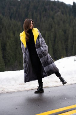 Kış dağlarında yolda poz veren bir kız. Gülümseyen, şık sarı ve gri uzun kürklü bir kadın. Kürk ve moda konsepti. Güzel insanlar..