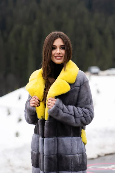 Κορίτσι που ποζάρει στο δρόμο σε χειμερινά βουνά φόντο. Λαμπερό αστείο νεαρή γυναίκα με χαμόγελο φορώντας κομψό κίτρινο και γκρι μακρύ γούνινο παλτό. Γούνα και μόδα έννοια. Όμορφοι άνθρωποι. — Φωτογραφία Αρχείου