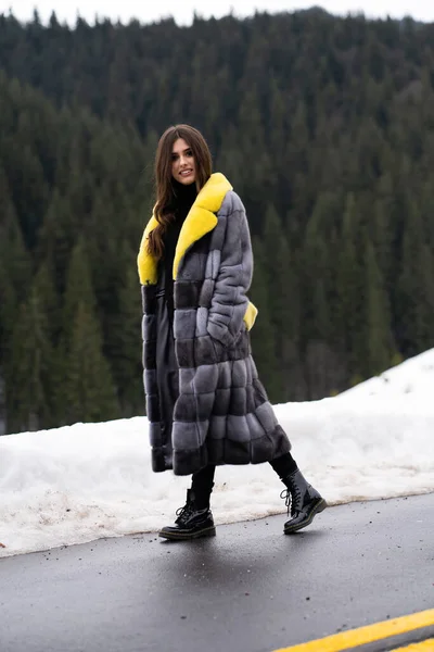 Девушка позирует на дороге на фоне зимних гор. Гламурная смешная молодая женщина с улыбкой в стильном желтом и сером длинном меховом пальто. Мех и концепция моды. Красивые люди . — стоковое фото