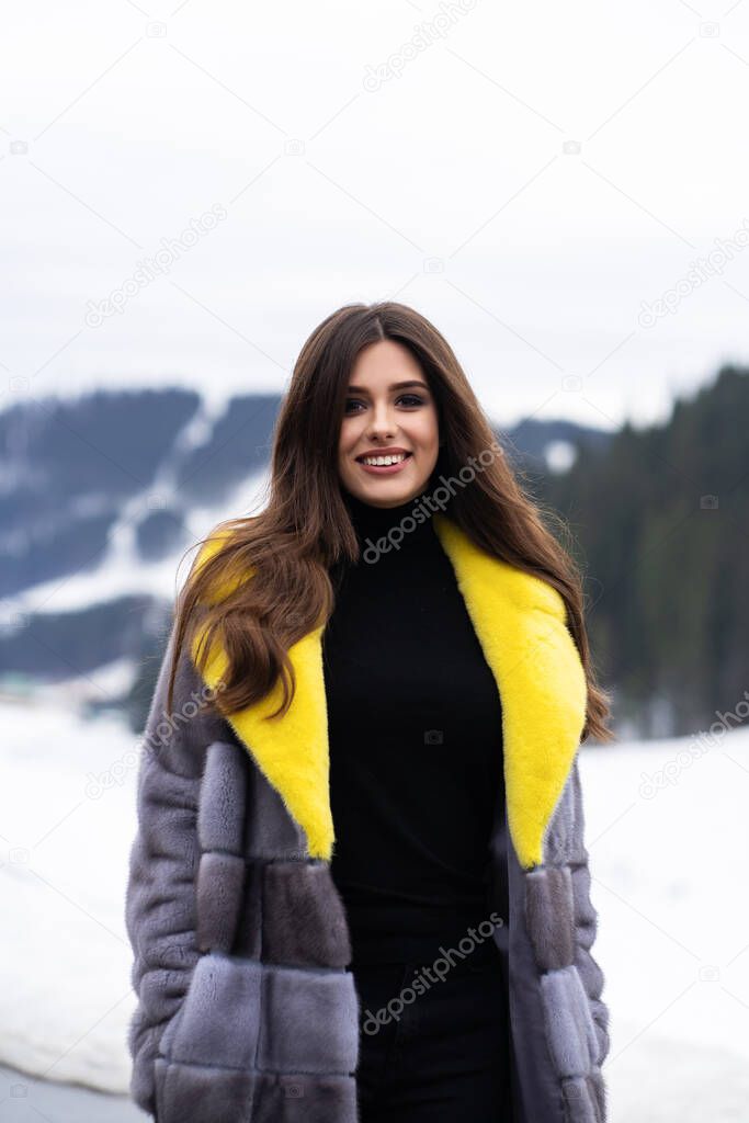 Girl posing on road on winter mountains background. Glamorous funny young woman with smile wearing stylish yellow and grey long fur coat . Fur and fashion concept. Beautiful people.
