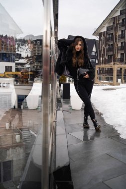 Kış dağlarında poz veren bir kız. Şık bir kazak, koyu kot pantolon, siyah moda kısa kürk ceket ve siyah çanta giyen alımlı genç bir kadın. Kürk ve moda konsepti. Güzel insanlar..