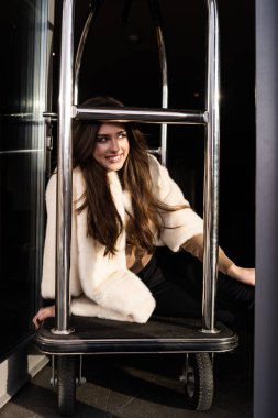 Seksi esmer kız otelin bagaj vagonuna oturur. Oteldeki bavullar. Şık beyaz kürk giyen çekici genç bir kadın. Moda, kürk, otel, seyahat ve güzel insanlar konsepti...