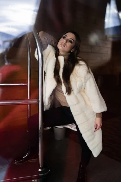 Σέξι μελαχρινή κοπέλα στέκεται κοντά στο καλάθι αποσκευών του ξενοδοχείου. Τρόλεϊ αποσκευών στο ξενοδοχείο. Λαμπερή νεαρή γυναίκα που φοράει κομψό λευκό γούνινο παλτό. Μόδα, γούνα, ξενοδοχείο, ταξίδια και όμορφα άτομα έννοια.. — Φωτογραφία Αρχείου