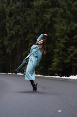 Kış dağlarının arka planında yola atlayan seksi esmer kız. Şık mavi uzun palto giyen alımlı genç bir kadın. Moda, iş ve güzel insanlar konsepti..