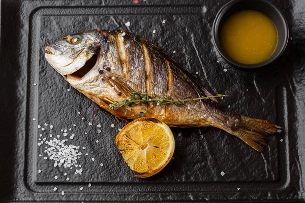 Delicious grilled dorado or sea bream fish with lemon slices, spices, rosemary on dark stone. Grilled sea fish with olive oil, spices and lemon ready for eating. Dorado, herbs and spices. Menu photo