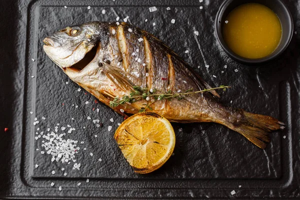 Delicious grilled dorado or sea bream fish with lemon slices, spices, rosemary on dark stone. Grilled sea fish with olive oil, spices and lemon ready for eating. Dorado, herbs and spices. Menu photo