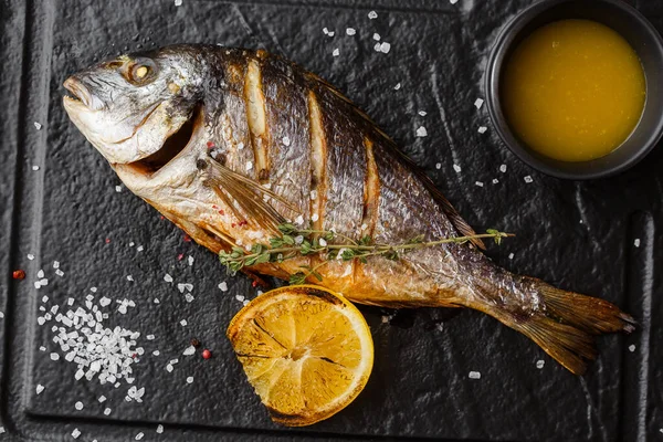 Delicious grilled dorado or sea bream fish with lemon slices, spices, rosemary on dark stone. Grilled sea fish with olive oil, spices and lemon ready for eating. Dorado, herbs and spices. Menu photo