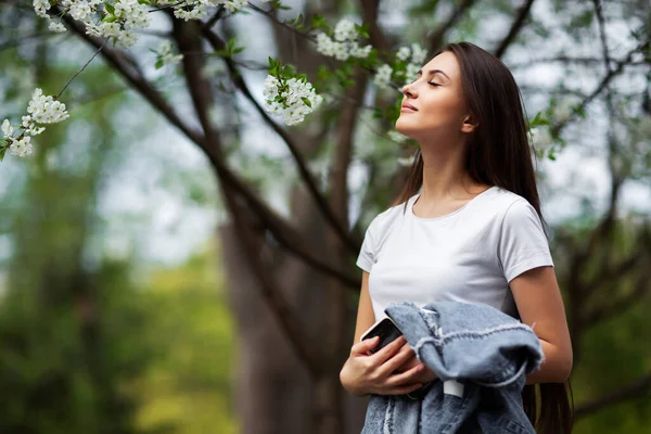 Doğal parktaki çiçek açan elma ağacının altında duran kız. Moda konsepti. Kot elbiseli zarif genç bayan güneşli bahar gününde çiçek açan bahçenin tadını çıkarıyor. Doğal güzellik makyajı. Yatay