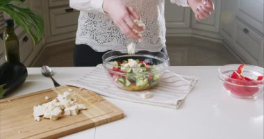 Kadın şeflerin elleri modern bir mutfakta tahta kesme tahtasına beyaz peynir döker. Kamera beyaz peynir koymayı takip ediyor. Salata hazırla. Sağlıklı yiyecekler. 6K Blackmagic kamerasıyla çekildi..