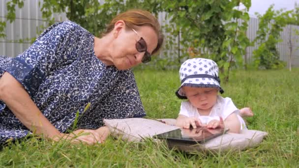 Kid with nanny watching cartoons. Cartoons on tablet watched outdoor. Girl lying on grass. Childhood, babyhood concept. Toddler with nanny outdoor. Grandmother with grandchild lying on green grass — ストック動画