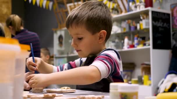 Kunstschule. Junge dekorieren Geschenk mit Pinsel. Zeichenstunde. Kunstwerkstatt. Kleiner Maler. Junge dekoriert Tonerzeugnis mit großem Interesse. Freizeitbeschäftigung für Kinder. Kind bereitet Geschenk vor. — Stockvideo