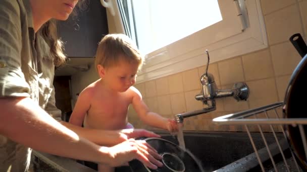 Anne bulaşıkları yıkarken çocuk lavaboda oturuyor. Anne ve kız kavramı. Mutfak görevleri. Ev işleri ve çocuklar. Çalışan anne ve yeni yürümeye başlayan çocuk. Anne elleri ve küçük tatlı kızı. — Stok video