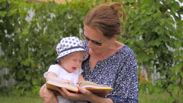 Reading concept. Woman holding book behind baby girl. Little girl looking into open book. Open book in womans hands. Toddler with mother reads the book. Close-up. American girl in panama — Stock Video