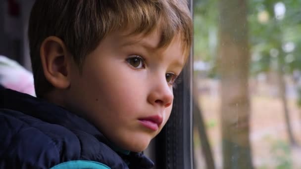 Smutny młody pasażer. Zbliżenie na małego ciekawskiego chłopca wyglądającego przez okno w pociągu. Wyraz twarzy. Zły dzień. Chłopak patrzy przez okno podczas podróży. Emocje twarzy małego podróżnika — Wideo stockowe