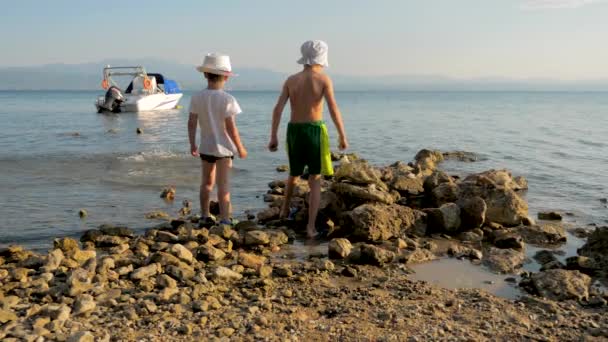 Két boldog kaukázusi kölyök, testvérek, együtt játszanak, köveket dobálva a tengerbe. Testvérek játszanak a szabadban kavicsos strandon a tenger ellen. Szabadság és boldogság fogalma — Stock videók