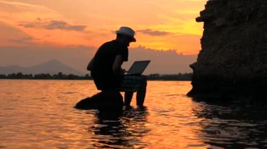 Happy man sitting on the stone and working at computer with sea view, static frame. Male silhouette on sunset seascape background. Travel blogger working on seaside. Freelancer concept.