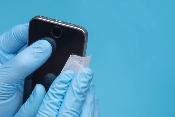 Hände in medizinischen Handschuhen wischen den Smartphone-Bildschirm mit Desinfektionstuch ab. Großaufnahme Hände reinigen Handy auf blauem Hintergrund. Nahaufnahme eines Arztes in Handschuhen, der sein Smartphone desinfiziert — Stockfoto