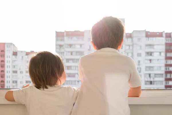 Copiii sunt forțați să fie acasă în timpul carantinei legate de pandemia de covid-19, frații se uită la fereastra deschisă a balconului. Carantină mondială, Coronovirus Pantemic, COVID-19 — Fotografie, imagine de stoc