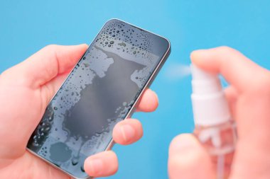 Close-up hands clean mobile phone on blue background. Closeup of a Doctor in gloves disinfecting his smartphone. Hands wiping mobile phone with alcohol decontaminating agent clipart