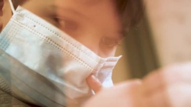 Yüz maskeli çocuk cep telefonuyla oyun oynuyor. Çocuk pencere eşiğinde oturmuş evde oyun oynuyor. Karantina sırasında ev okulunda. Pencereden bak. Önleme salgını