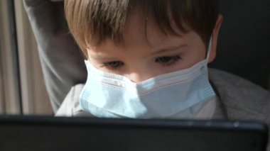 Tıbbi maske takan çocuğun yüzünü kapatın. Evde kalıp koronavirüs salgınını önleyin. Koruyucu tıbbi maskeli çocuk pencere pervazına oturur ve oyun oynar. Pencereden bak. Dizinin bir parçası
