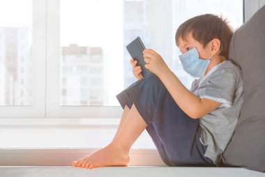 Çocuk evde tablet kullanarak online çalışıyor. Küresel salgın covid19, Coronavirus. Koruyucu tıbbi maskeli çocuk pencere pervazına oturur ve oyun oynar. Ev konseptinde kal. Evde öğrenim.