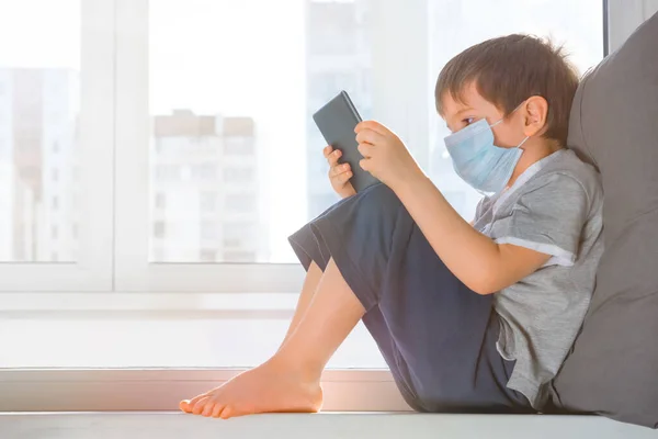 Хлопчик вчиться вдома, використовуючи планшет. Global pandemic covid19, Coronavirus Дитина в захисній медичній масці сидить на підвіконні і грає в ігри. Залишайся вдома з концепцією. Навчання вдома. — стокове фото