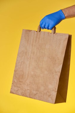 Mavi eldiven sarı arka planda kağıt torba tutuyor.