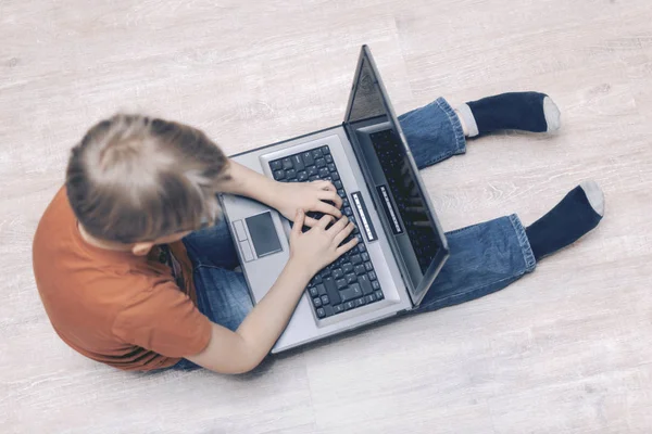 Ein Laptop auf den Füßen eines Jungen, der auf dem Boden sitzt. Die Hände sind auf der Tastatur. Computerspiele spielen, trainieren, bloggen, tippen. Ansicht von oben. Selektiver Fokus — Stockfoto