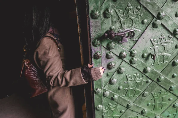 Palto giymiş bir kadın figürü ve elinde sırt çantası olan eski demir bir kapı.