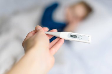 Hand with thermometer in hand, taking temperature, and recovering child on the background. Virus, contagious disease, coronovirus concept.