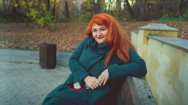 Um retrato de uma bela mulher adulta ruiva, sentado em um banco perto da vegetação. — Fotografia de Stock