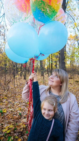 Eine junge Mutter und ihre kleine entzückende Tochter mit den bunten Luftballons spazieren im herbstlichen Wald. — Stockfoto