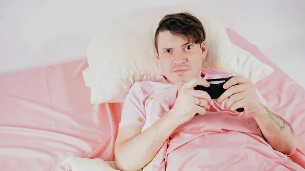 Hombre con joystick jugando a videojuegos acostado en la cama. Un hombre joven con un joystick, concepto de adicción a los juegos, ocio en casa — Foto de Stock