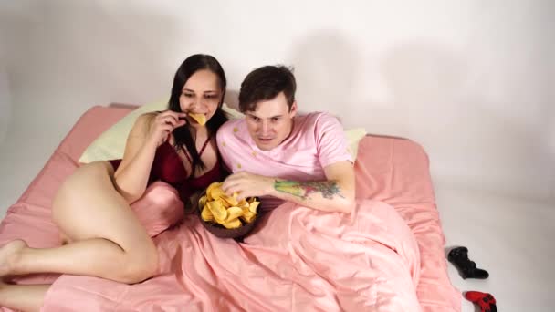 Una giovane coppia sposata si trova a letto, guarda un film e mangia patatine fritte. La donna e l'uomo ridono di un divertente film comico. — Video Stock